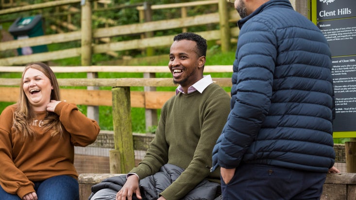 A close-up of two visitors sitting on a wall and laughing, with another visitor standing in front of them at Clent Hills